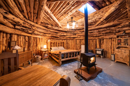 Cozy wooden cabin interior with a bed, stove, and rustic furniture, featuring a skylight and log walls.